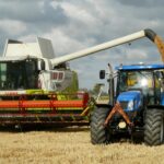 A modern combine harvester and tractor working together to harvest wheat in a sunny outdoor field.